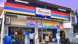 Bagian depan gerai indomaret terdapat pegawai indomaret dan pembeli dengan anak kecil.